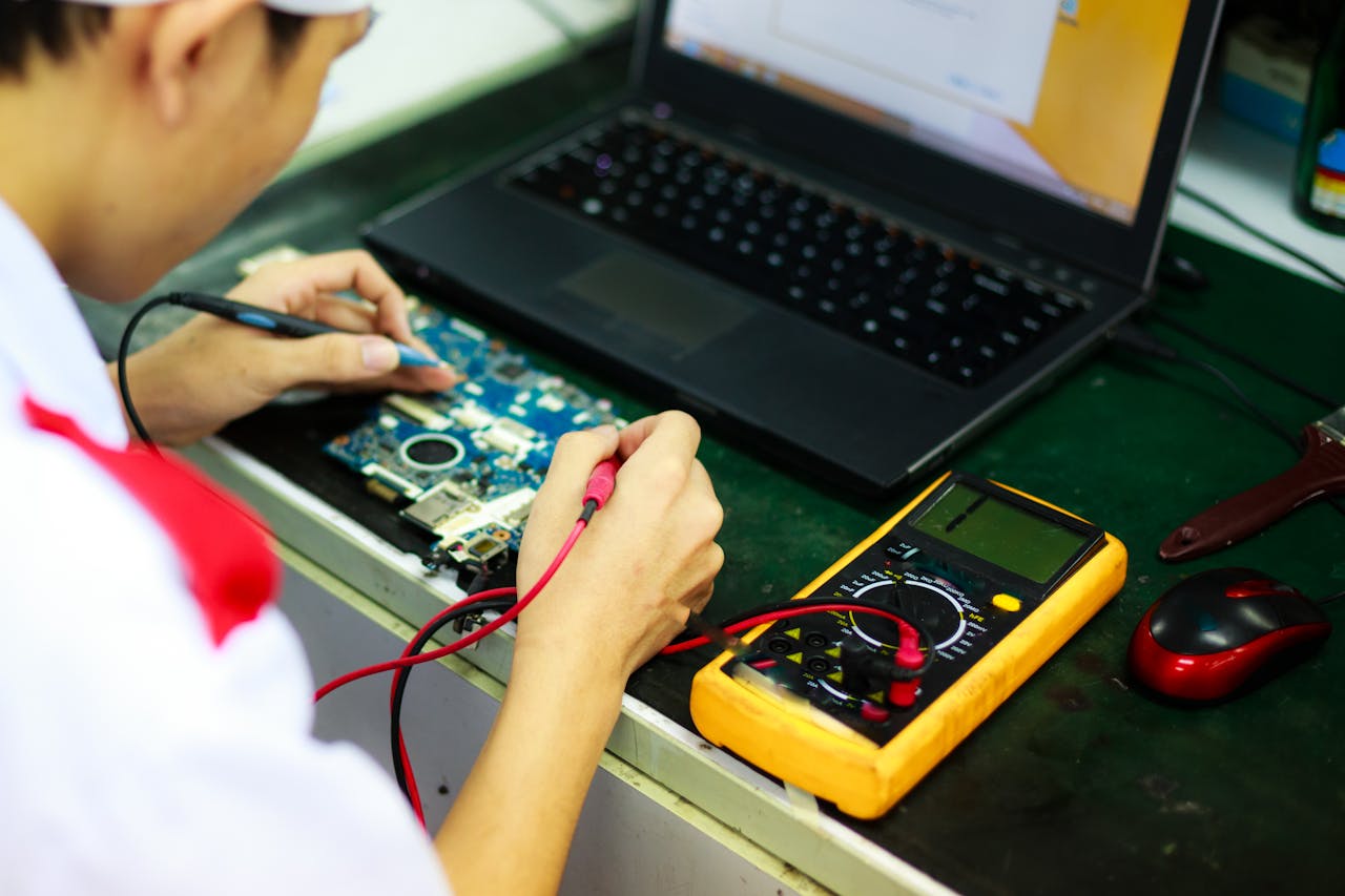 Services Technician working on a laptop with multimeter in a professional workspace.